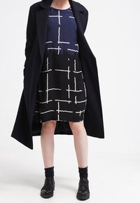 Musta takki vaaleansinisessä mekossa, jossa on valkoinen abstrakti kuvio. Mekossa on lyhyet hihat ja pyöreä kaula-aukko. Mustat nilkkasaappaat ja sukat.