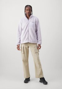 Hele lilla fleecejope esiküljel lukuga ja rinnataskuga, kombineeritud heledate beežide cargo-pükstega ja mustade tennistega.