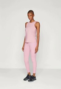 Ženska stoji v roza brez rokavov in leggingah ter črnih športnih čevljih pred enobarvno svetlo ozadje.