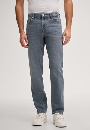 Mężczyzna noszący szare jeansy o prostym kroju i białe trampki, stojący na tle jasnego, jednolitego tła.