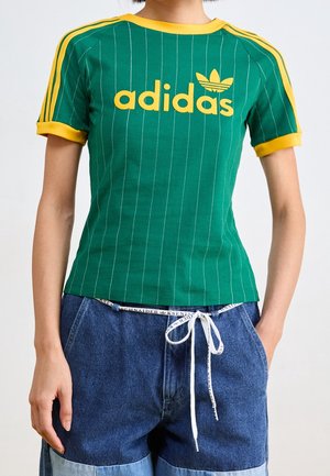 Persona con una camiseta verde de Adidas con rayas y logo amarillos, combinada con pantalones cortos de mezclilla azul de dos tonos con un cinturón de cuerda blanco.