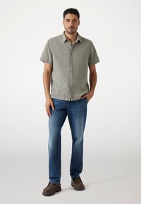 Camisa de botones de manga corta en verde claro con una textura sutil, combinada con jeans azules de lavado medio y calzado marrón.
