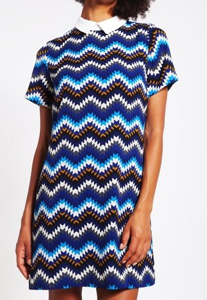 Frau trägt ein kurzärmeliges, knielanges Kleid mit weißem Kragen, das ein blaues, schwarzes, weißes und braunes Zickzack-Geometriemuster zeigt.