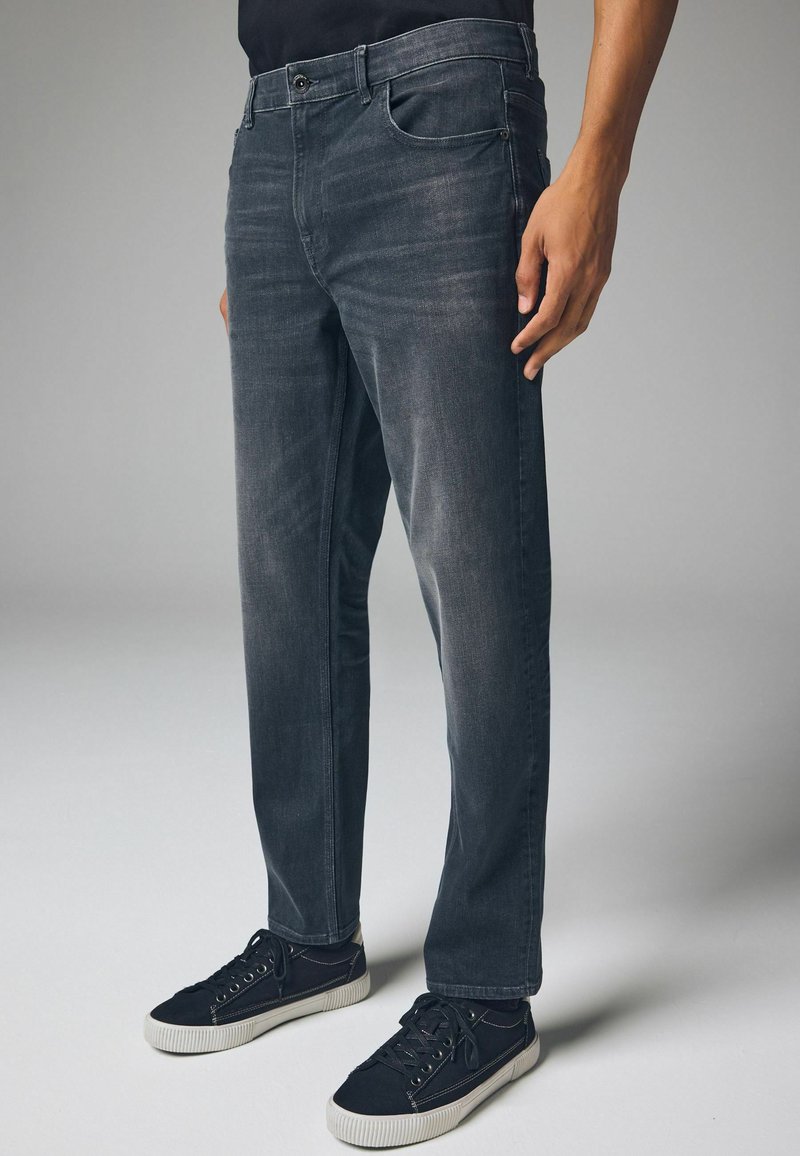 Donkergrijze spijkerbroek van denim, met een standaard pasvorm, vijf zakken en een subtiele vervaging. Ggedragen met zwarte sneakers.