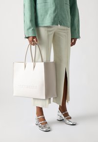 Sac fourre-tout en cuir beige avec logo "Coccinelle" en relief, texture lisse et deux poignées. Porté avec une veste vert menthe et un pantalon clair.