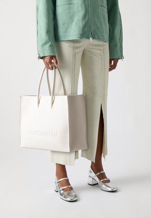 Geantă tote din piele bej cu logo-ul embosat "Coccinelle", textură netedă și două mânere. Purtați cu o jachetă verde mentă și pantaloni deschiși.