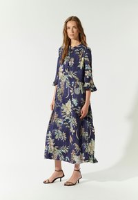 Maxikleid mit floralem Druck in Marineblau, 3/4-Ärmeln, Rüschenmanschetten und schwingendem Rock. Kombiniert mit schwarzen, geschnürten hohen Schuhen.