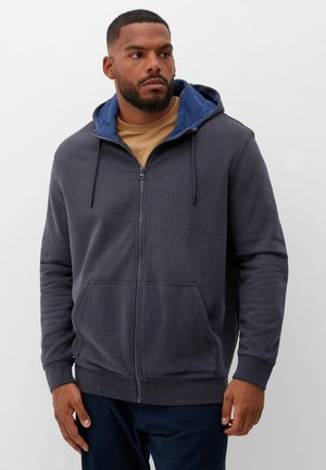 Mann mit Bart, der einen dunkelgrauen Kapuzenpullover mit Reißverschluss über einem hellbraunen Hemd trägt, steht vor einem einfarbigen hellen Hintergrund und schaut nach links.