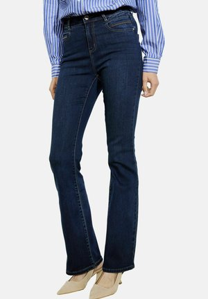 Jeans a zampa d'elefante in denim blu scuro con vita media, caratterizzati da cinque tasche, cuciture a contrasto e una leggera lucentezza, abbinati a tacchi beige.