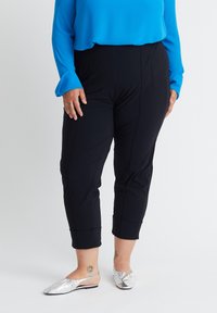 Schwarze, taillierte Hosen mit Falten, ausgestattet mit Seitentaschen und einer glatten Textur. Kombiniert mit einer leuchtend blauen Bluse und silbernen Slippern.