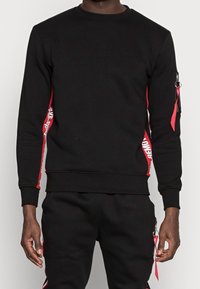 Man som bär en svart sweatshirt och byxor med röda sidostråk, med vit text och röda dragkedjesnoddar på vänster arm och byxfickan.