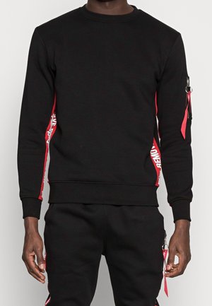 Man in een zwart sweatshirt en broek met rode zijsstripes, voorzien van witte tekst en rode rits trekt aan de linkerarm en broekzak.