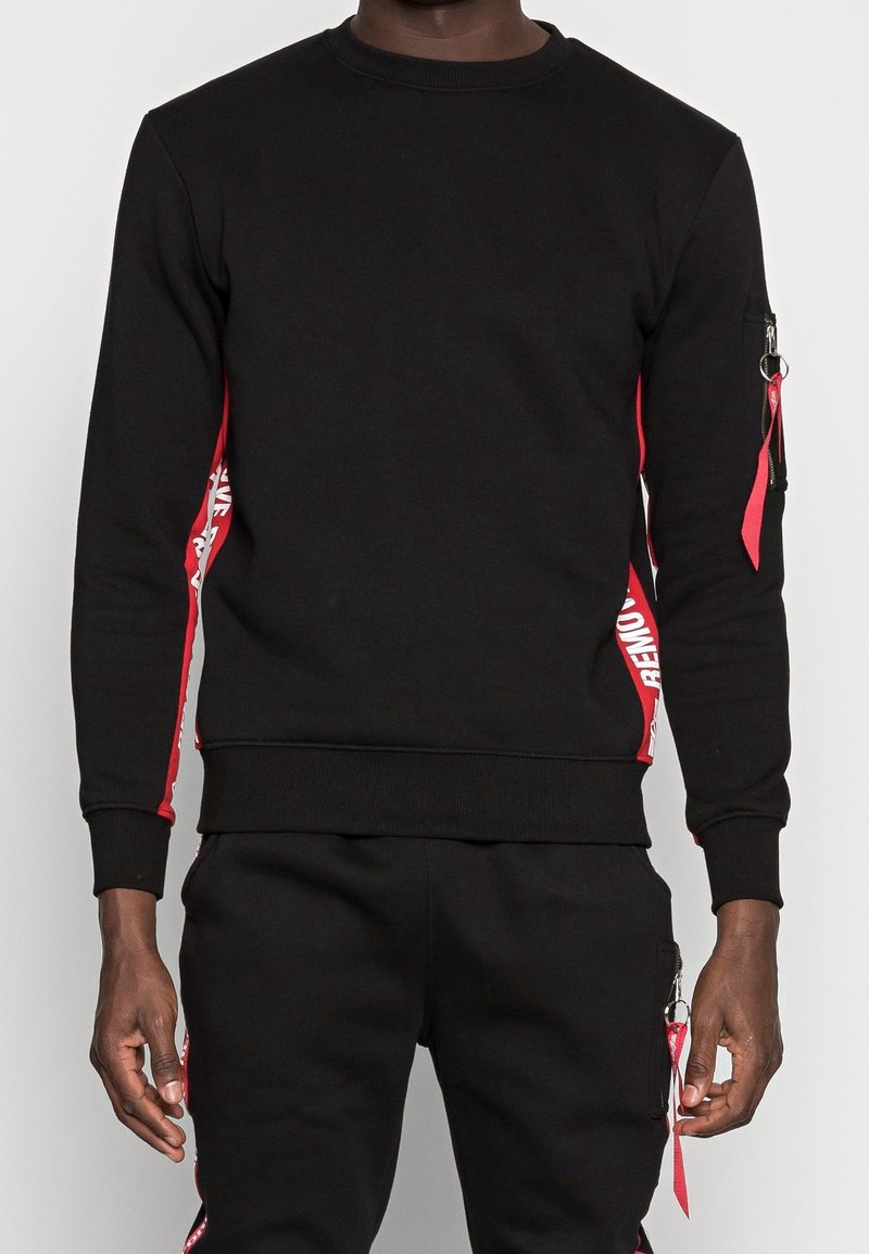 Man som bär en svart sweatshirt och byxor med röda sidostråk, med vit text och röda dragkedjesnoddar på vänster arm och byxfickan.