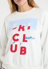 Biała bluza z napisem "SKI CLUB" na czerwono, z grafiką gór i narciarza w kolorze niebieskim. Żebrowany dekolt i dół, miękka teksturowana tkanina.