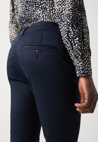 Gros plan d'une personne portant un pantalon bleu marine avec une poche arrière à bouton et un chemisier à manches longues à motifs tachetés avec manches à poignets.