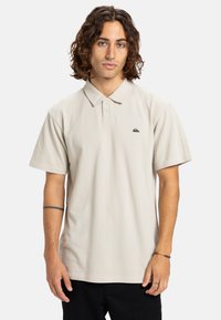 Lys beige polo shirt med klassisk krave, korte ærmer, tre-knaps lukning og et lille sort logo på brystet. Blød tekstur, afslappet pasform.