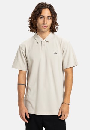 Light beige polo shirt with a classic collar, short sleeves, three-button placket, and a small black logo on the chest. Soft texture, casual fit.