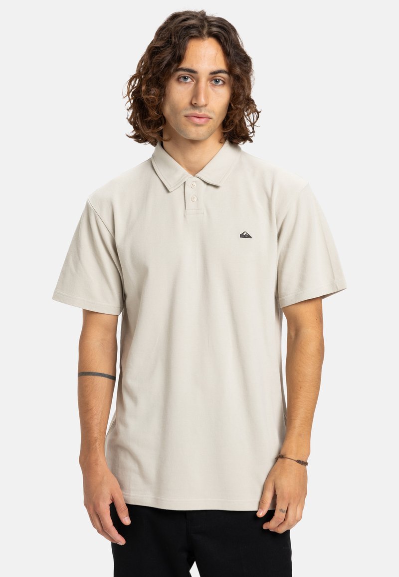 Lys beige polo shirt med klassisk krave, korte ærmer, tre-knaps lukning og et lille sort logo på brystet. Blød tekstur, afslappet pasform.