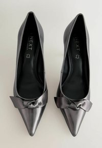 Schwarze Lackleder-Highheels mit einer spitzen Zehenkappe und einem auffälligen Schleifendetail auf dem Spann, das eine glatte, reflektierende Oberfläche aufweist.