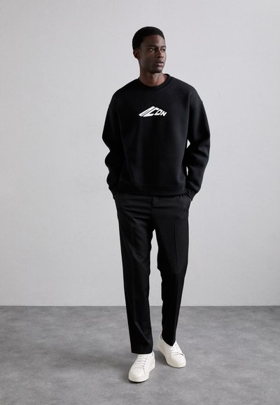 Felpa nera con logo bianco, abbinata a pantaloni neri e sneakers bianche. Tessuto morbido, vestibilità rilassata e design semplice.