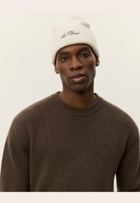Stickad mössa i krämfärg med uppvikt kant och broderad logotyp. Modellen bär en brun texturerad tröja med ribbat mönster.