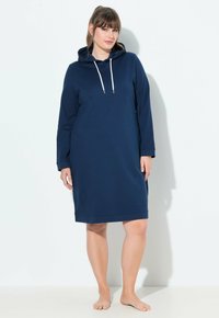 Donna in piedi a piedi nudi che indossa un vestito a felpa blu navy con cordini bianchi su uno sfondo bianco semplice.