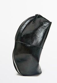 Sort taske i laklæder med en krøllet tekstur, buet form og en markant sølvlynlås langs kanten.