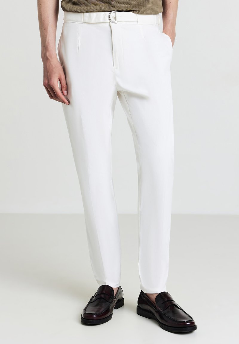 Homme portant un pantalon blanc slim-fit avec une ceinture et des mocassins en cuir marron foncé, se tenant devant un fond clair et uni.