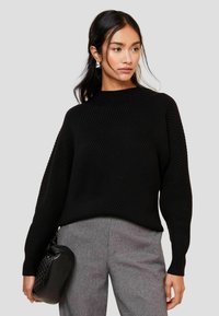 Schwarzer gerippter Pullover mit hohem Kragen und weiten Ärmeln, kombiniert mit grauen Hosen und einer strukturierten schwarzen Handtasche.