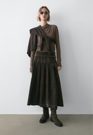 Brązowa bluzka z długim rękawem połączona z ciemną plisowaną spódnicą. Modelka ma na sobie ciemne owalne okulary przeciwsłoneczne i zielone, sięgające kostek buty.