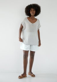 Beżowo-białe, prążkowane sweter w luźnym kroju z dekoltem w serek. Połączony z jasnymi szortami i płaskimi sandałami, na neutralnym tle.