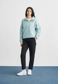 Νεαρή γυναίκα που στέκεται με το χέρι στην τσέπη, φοράει ανοιχτό μπλε φλις μπουφάν, μαύρο παντελόνι, λευκές κάλτσες και λευκά αθλητικά παπούτσια σε μπλε πάτωμα.