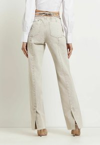 Jeans acampanados beige de cintura alta, con dos bolsillos traseros, costuras divididas en los tobillos y bordes deshilachados. Llevados con un top corto blanco.