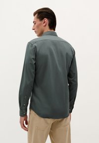 Camisa de manga larga verde oscuro con puños abotonados y un ligero brillo. Presenta un cuello clásico y un dobladillo curvado, combinada con pantalones beige.