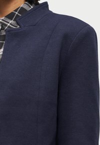 Κοντινό πλάνο ενός ατόμου που φοράει ένα μπλε σακάκι με υφή πάνω από το γιακά ενός ασπρόμαυρου πουκαμίσου με καρό σχέδιο.