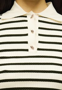 Πλεκτό πόλο πουκάμισο με οριζόντιες ρίγες σε κρεμ και σκούρο πράσινο, με επίπεδη γιακά και τέσσερα διακοσμητικά κουμπιά με μοτίβα φύλλων.