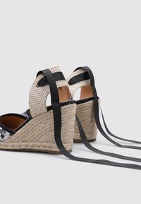 Sandalias de cuña con un tacón de yute trenzado, parte superior de textil negro con un diseño texturizado y correas de tobillo tejidas en beige aseguradas con cintas negras.
