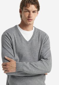 Grauer V-Ausschnitt-Pullover aus weichem Strickmaterial, mit gerippten Bündchen und klassischem Design. Über einem weißen T-Shirt getragen.