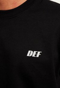 Sort crewneck sweatshirt lavet af bomuld. Har et lille hvidt logo "DEF" på venstre side af brystet. Glat tekstur, afslappet stil.