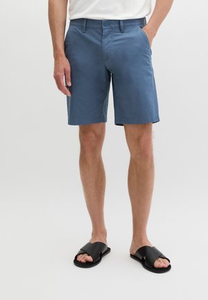 Mann trägt knielange blaue Shorts und schwarze Slide-Sandalen, steht mit einer Hand in der Tasche vor einem schlichten weißen Hintergrund.