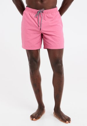 Man in felroze zwembroek met zwart-witte trekkoord, staand op blote voeten tegen een effen lichte achtergrond.