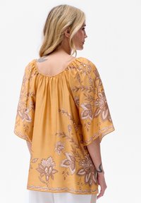 Krótki rękaw musztardowa bluzka z kwiatowym haftem, zebranym dekoltem i swobodnym krojem. Posiada misternie wykonane brązowe kwiatowe wzory.