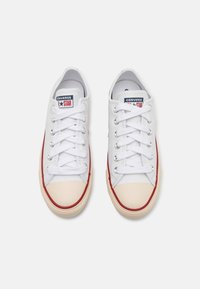 White canvas sneakers with a red stripe, rounded beige toe cap, flat laces, and logo patches on the tongue. Rubber sole with textured edge.