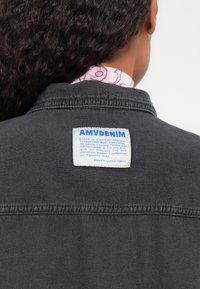 Čierna denimová bunda s výraznou bielou nálepkou, na ktorej je nápis "AMV DENIM." Medzi vlastnosti patrí klasický golier a viditeľné švy na lemovaní.