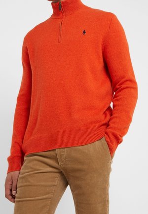 Mężczyzna ubrany w jaskrawo pomarańczowy sweter z suwakiem do połowy długości i ściągaczami na rękawach oraz beżowe spodnie z sztruksu, z jedną ręką w kieszeni.