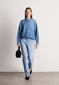 Denim srajca in svetlo modri kavbojki, oba v sproščenem kroju; temna torbica v roki; nizki črni čevlji; gladek material in svetel pranje.