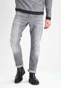 Jean en denim gris avec une coupe slim, présentant une texture délavée et des ourlets retroussés, portés avec des bottines noires.