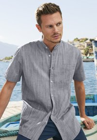 Korte mouwen button-up shirt van lichtgrijze gestreepte stof, met een bandkraag en een borstzak, weergegeven tegen een schilderachtige waterkant achtergrond.