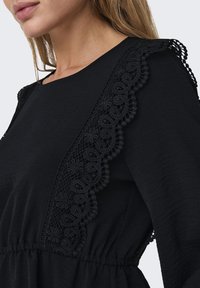 Svart långärmad klänning med intrikata spetsdetaljer längs axlarna, texturerad tyg och ett elastiskt midjeband för att ge form.