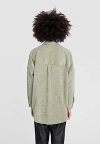 Persona con capelli ricci che indossa una maglietta a maniche lunghe verde chiaro e pantaloni neri, in piedi rivolto di spalle contro uno sfondo semplice.